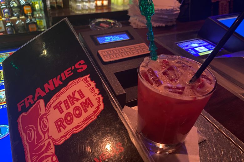 A red cocktail with a green tiki stirrer sits on a bar next to a Frankie's Tiki Room menu and a video poker machine.