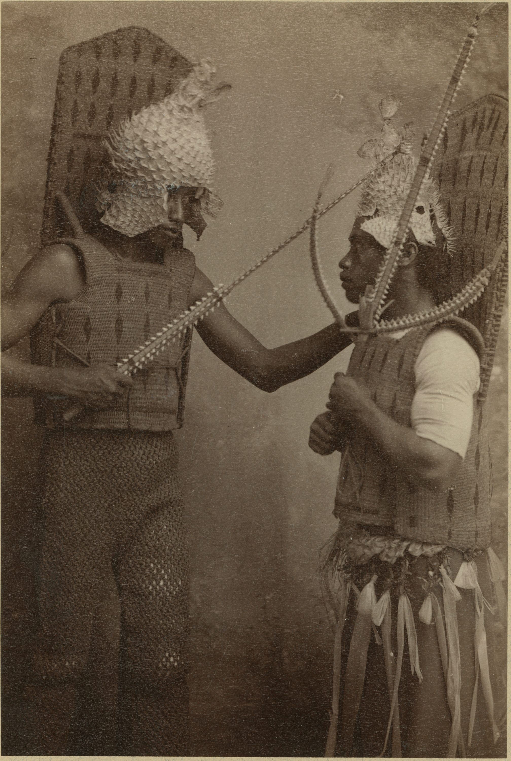 Two men with porcupine fish helmets, coconut fiber armor, and <em>taumangaria</em>. 