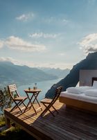 A bed with white fluffy bedding on a wooden open-air platform overlooking a river and mountains.