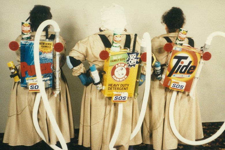 Three people in tan coats wear homemade Ghostbusters-style proton packs made from laundry detergent boxes and hoses.