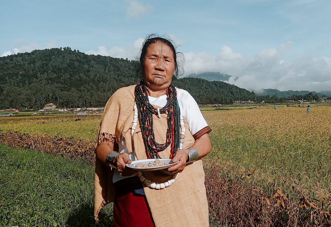 An elder Apatani woman holds a plate of tapyo in the paddy fields.