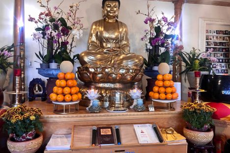 A buddhist altar with a golden seated buddha flanked by flowers and oranges.