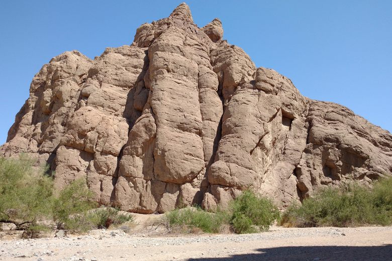 A large, craggy brown rock formation rises above desert shrubs and a sandy ground under a clear blue sky.