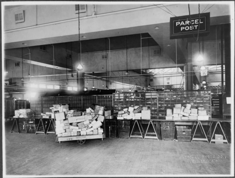 Parcel post area of mail room showing trucks and tables stacked with packages, U.S. Post Office, Washington, D.C, c. 1920