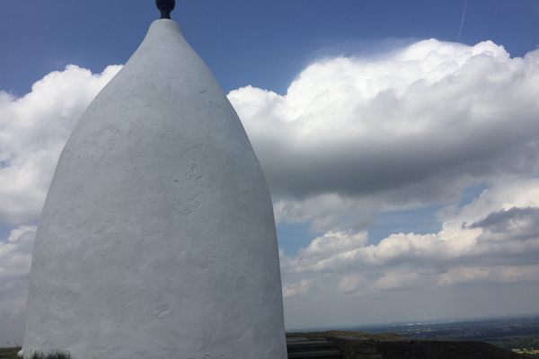 White conical shaped structure with a black bulb-like pinnacle with clouds in the background.