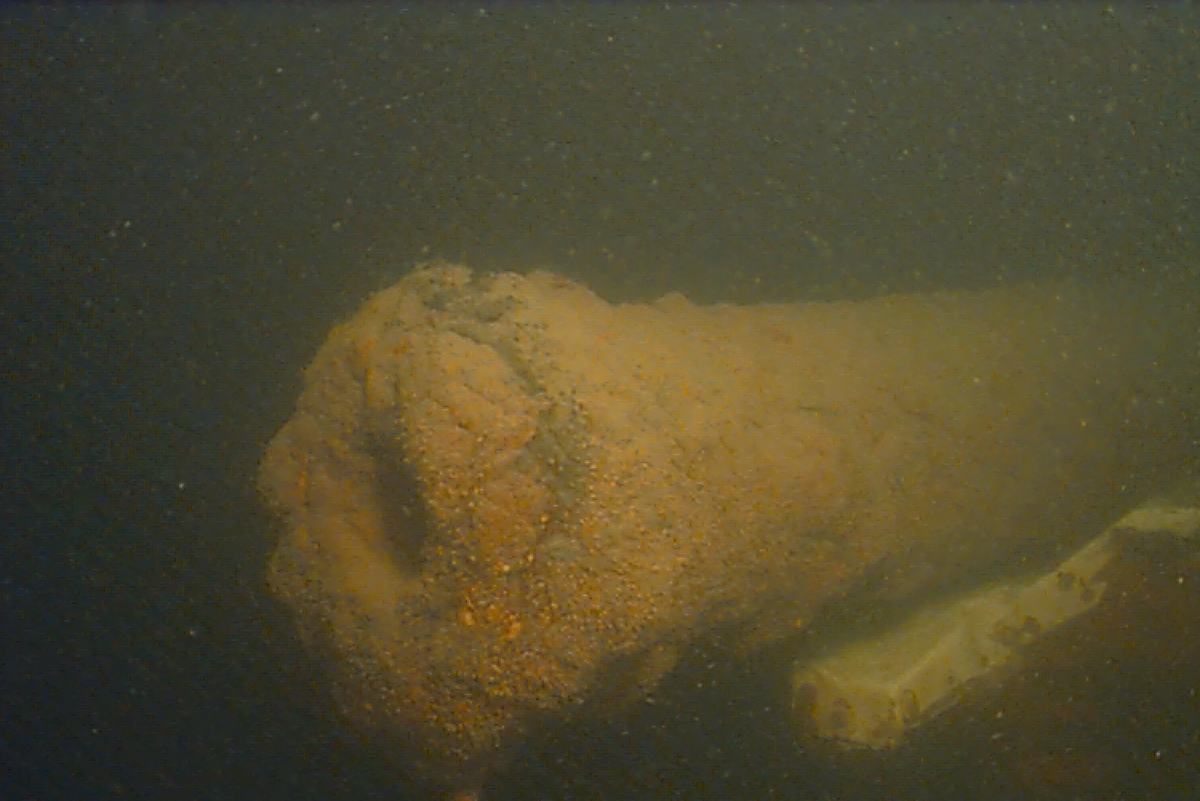 The <em>Spitfire</em>'s bow cannon is covered by a concretion of rust and sediment, formed over centuries in the underwater environment. The piece of modern metal in lower right of image is part of a sign stating the wreck is protected under the Sunken Military Craft Act, making any unauthorized activity at the site, including artifact recovery, punishable by law.