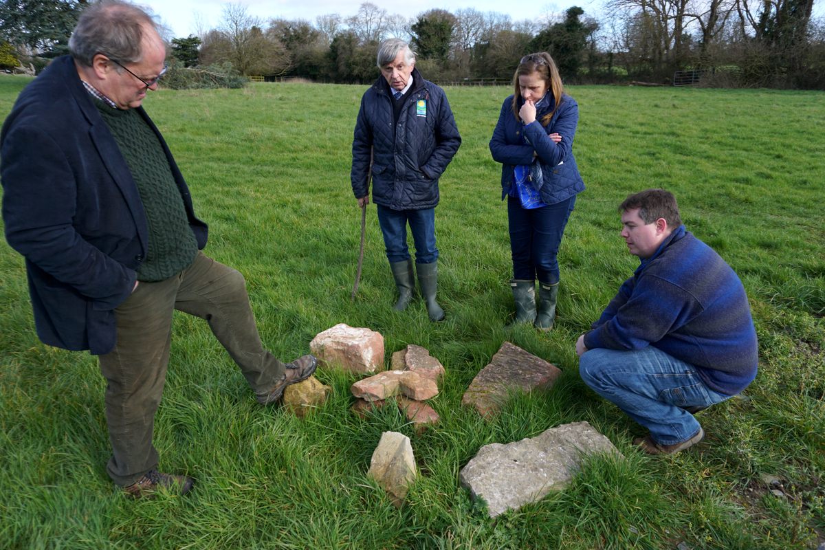 Archaeologist Matthew Stout rests his foot on a piece of red sandstone, a clue to the site's builders, as he discusses the site with landowner John McCullen, his daughter Grace McCullen, and conservationist Brandon Good.