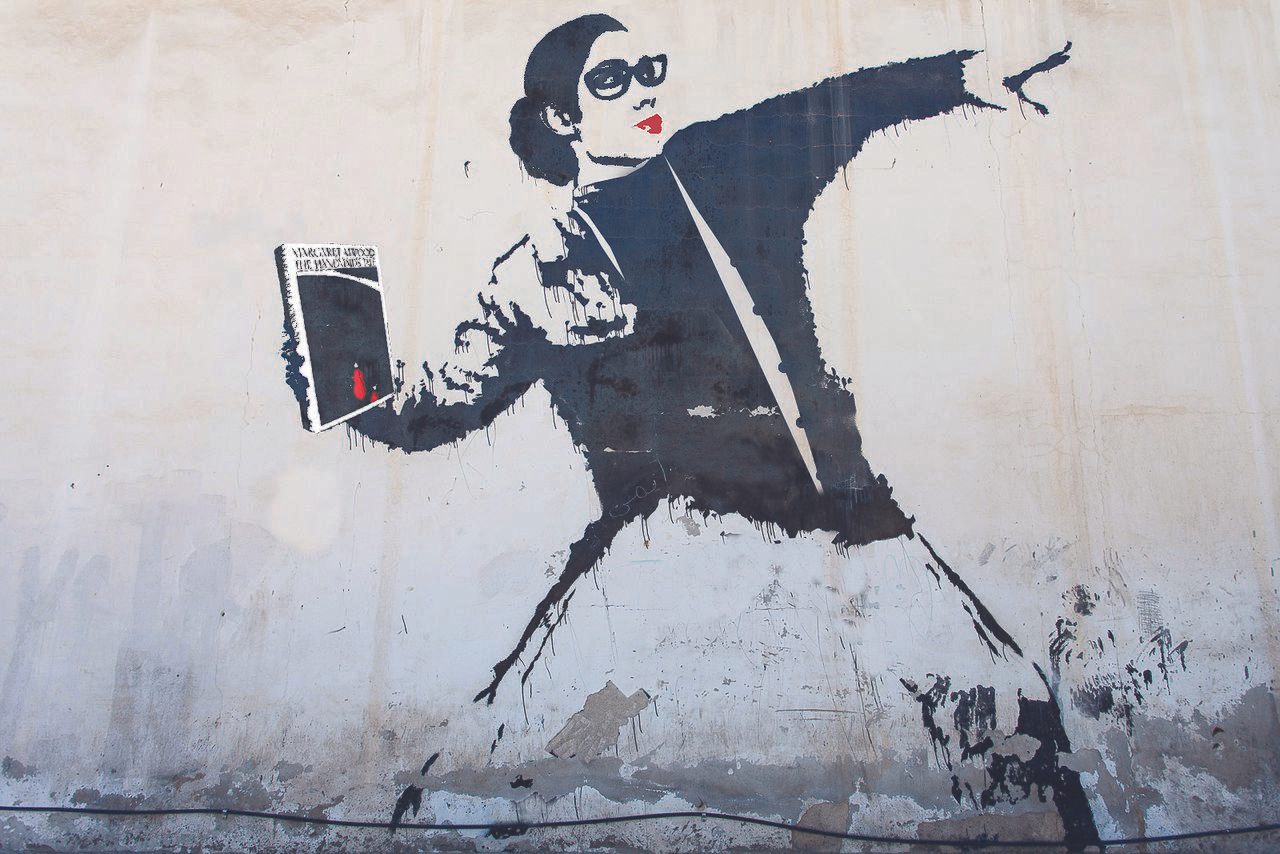 An adaptation of Banksy's "Flower Bomber," this depicts a librarian in protest, throwing Margaret Atwood's <em>A Handmaid's Tale</em>.
