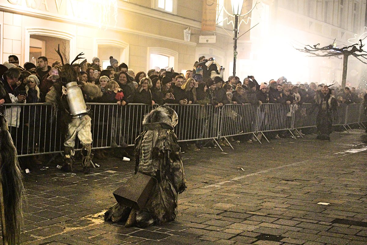 The crouching Krampus is pausing dramatically before leaping over a barrier into the crowd. The standing Krampus is beating an onlooker with a birch whip, and he has several spare weapons on his back.