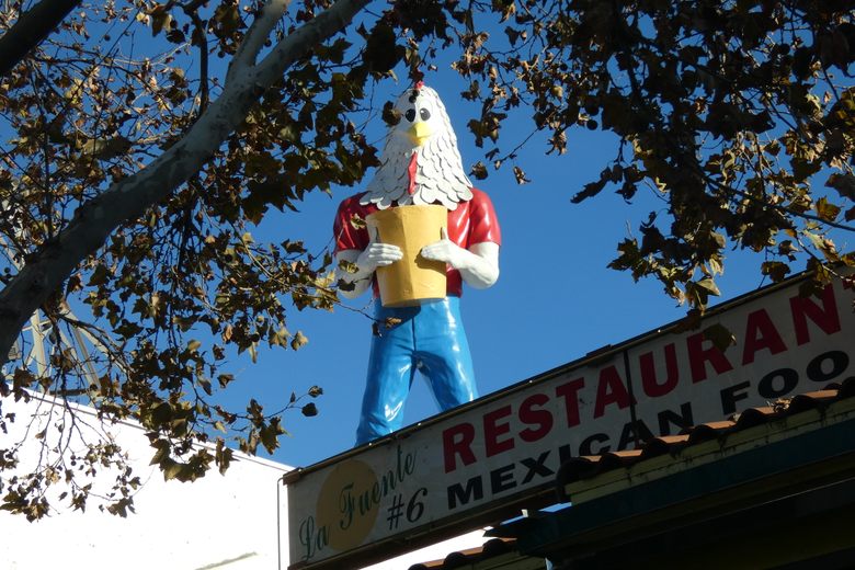 A tall fiberglass statue of a man with a chicken head holding a yellow bucket atop a restaurant roof.