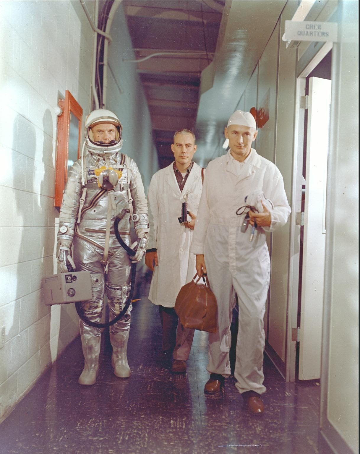 Astronaut John H. Glenn Jr., NASA flight surgeon William Douglas and equipment specialist Joseph W. Schmidt leave crew quarters prior to the Mercury-Atlas 6 mission. Glenn is in his pressure suit and is carrying the portable ventilation unit.