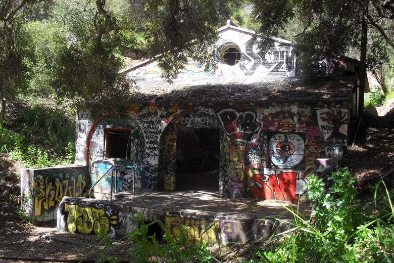A small, abandoned stone building covered in colorful graffiti sits nestled among trees in a wooded canyon.