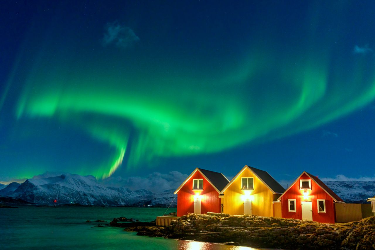 Aurora Borealis over snowcapped mountains and fishermen cabins by the sea in Sommaroy, Troms og Finnmark, Norway.
