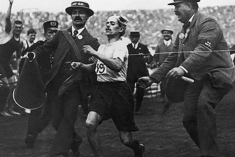 Dorando Pietri is helped across the finish line while holding a cork in his hand. 