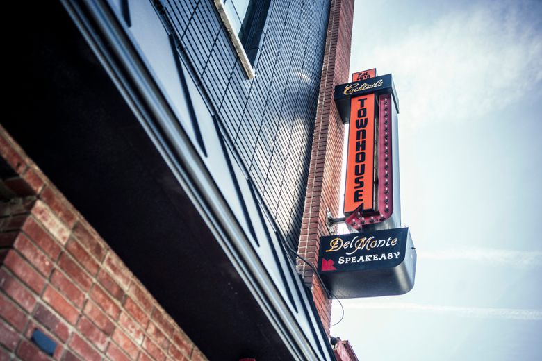 A vertical neon sign for Townhouse and Del Monte Speakeasy hangs on a brick building against a pale blue sky.