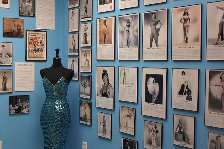 A blue wall at the Burlesque Hall of Fame covered in framed vintage photos of performers beside a sequined gown.