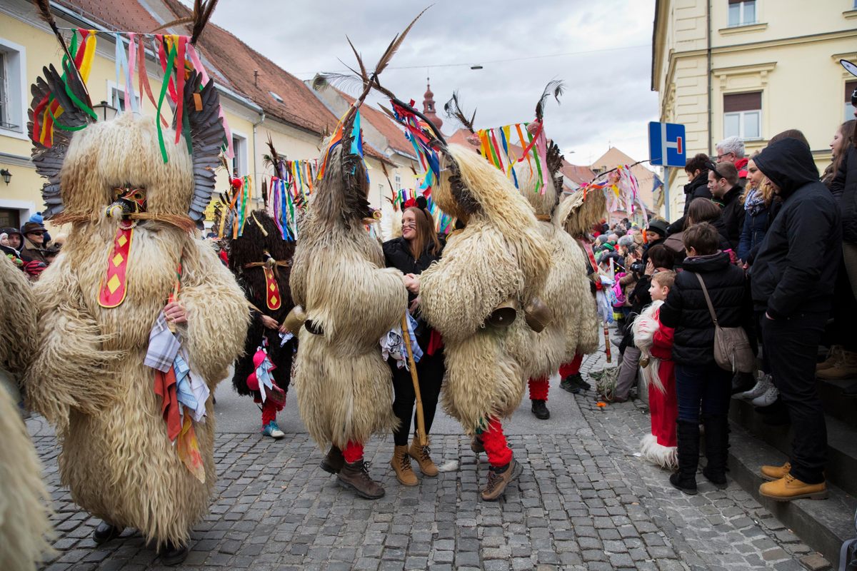 Two Kurenti surround a woman. After they jump around her, she will tie a handkerchief on their <em>jezevka</em>, the wooden bats they carry studded with hedgehog spikes.