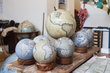 Mini Desk Globes and an Ochre Livingstone desk globe in the studio. The company uses a variety of wood and metal to make their bases. Here you can see Zebrano, solid Copper, Walnut, solid Aluminium. The wood bases are hand turned and the metal ones are hand cast using the lost wax method. All made by hand and finished by hand here in London. On the desk also sits a practice globe made by an apprentice that has not yet been stripped (The two tone one). Photo by Ana Santl.