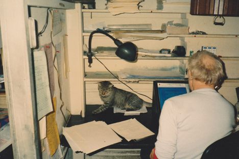 An unidentified Studio Duplicating employee uses a homemade, oversized magnifying glass to compare a script typed in mimeostencil to the original manuscript to his left, assisted by the office cat.