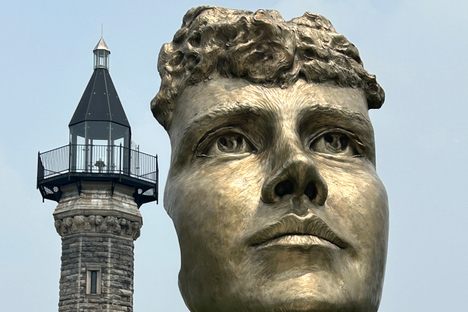 A bronze fast of a female face atop a pedestal with a lighthouse in the background