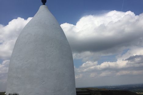White conical shaped structure with a black bulb-like pinnacle with clouds in the background.