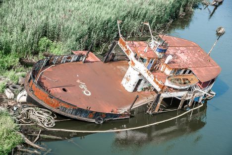 A rusty ship sits on a grassy riverbank.