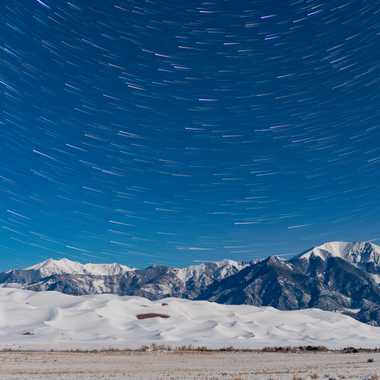 Star trails trace the night sky above snow-covered mountains and sand dunes.