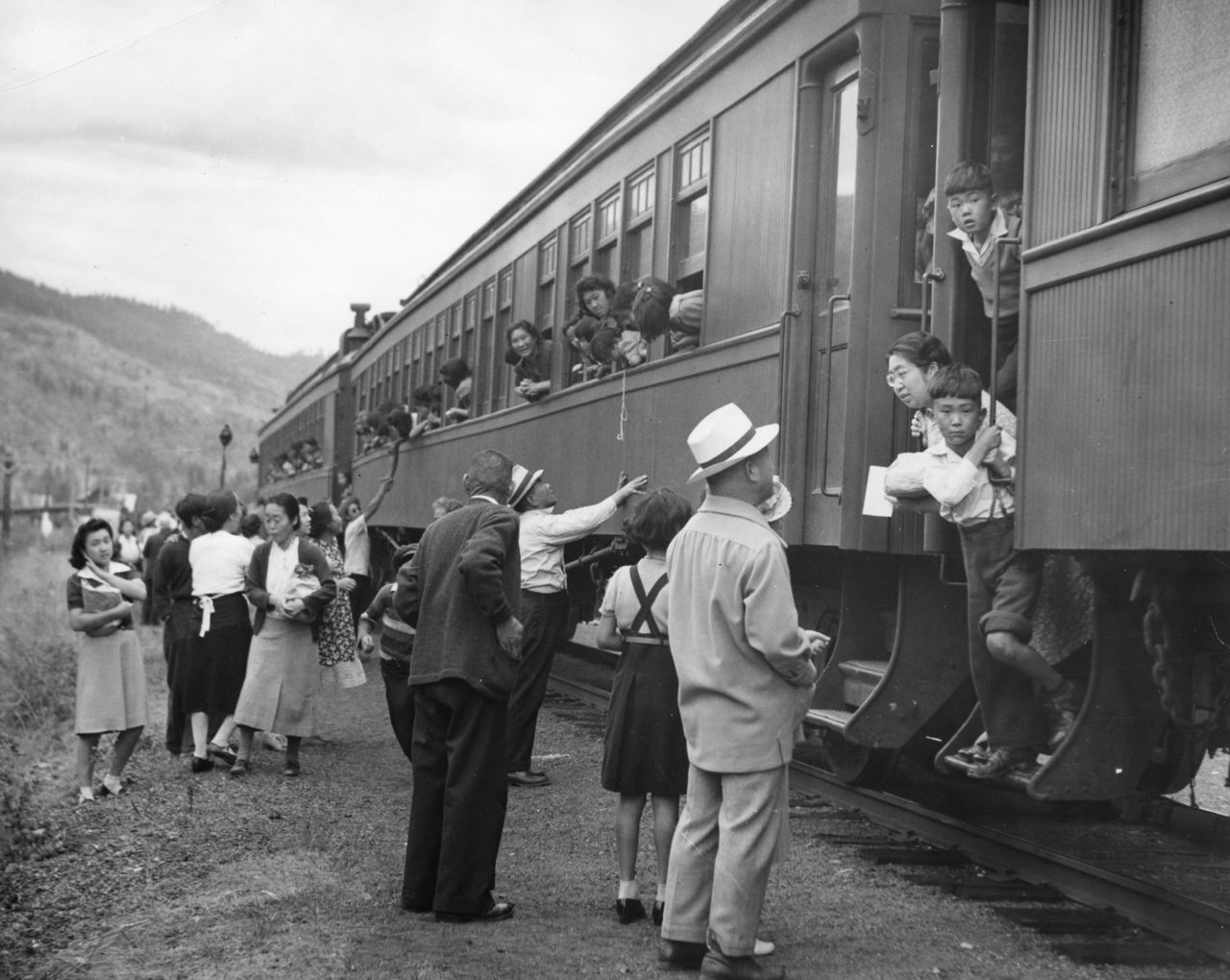 Displaced Japanese Canadians leaving the Vancouver area (possibly Slocan Valley) after being prohibited by law from entering a “protected area” within 100 miles of the coast. The woman on the left in this Leonard Frank photograph holding the book is Nobuko Morimoto, the woman in dark cardigan is Tei Terashita, and the young woman leaning out of train is identified as Kazuyo Kawabata.