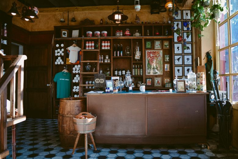 A wooden shop counter sits before floor-to-ceiling shelves stocked with bottled goods, books, and quirky merchandise.