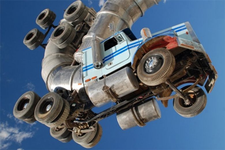 Two semi-trucks are intertwined and curved into a vertical S-shape against a clear blue sky.
