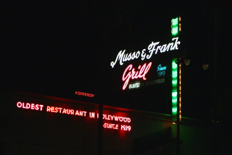 The glowing neon signs of Musso and Frank Grill stand out against a dark night sky in Hollywood.