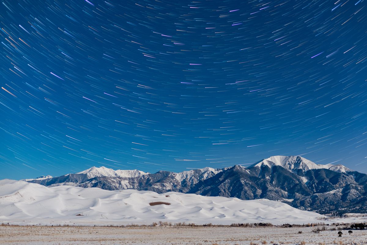Star trails trace the night sky above snow-covered mountains and sand dunes.