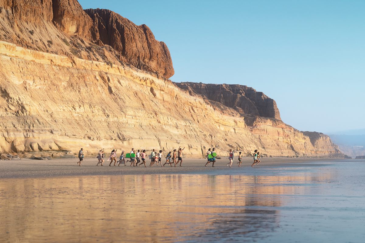 On a California beach with big cliffs in the background, a group of people of all ages head towards the ocean with boogie boards in hand.