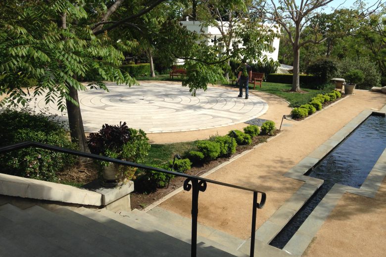 A person walks on a circular stone labyrinth in a lush garden with a long reflecting pool and leafy trees.