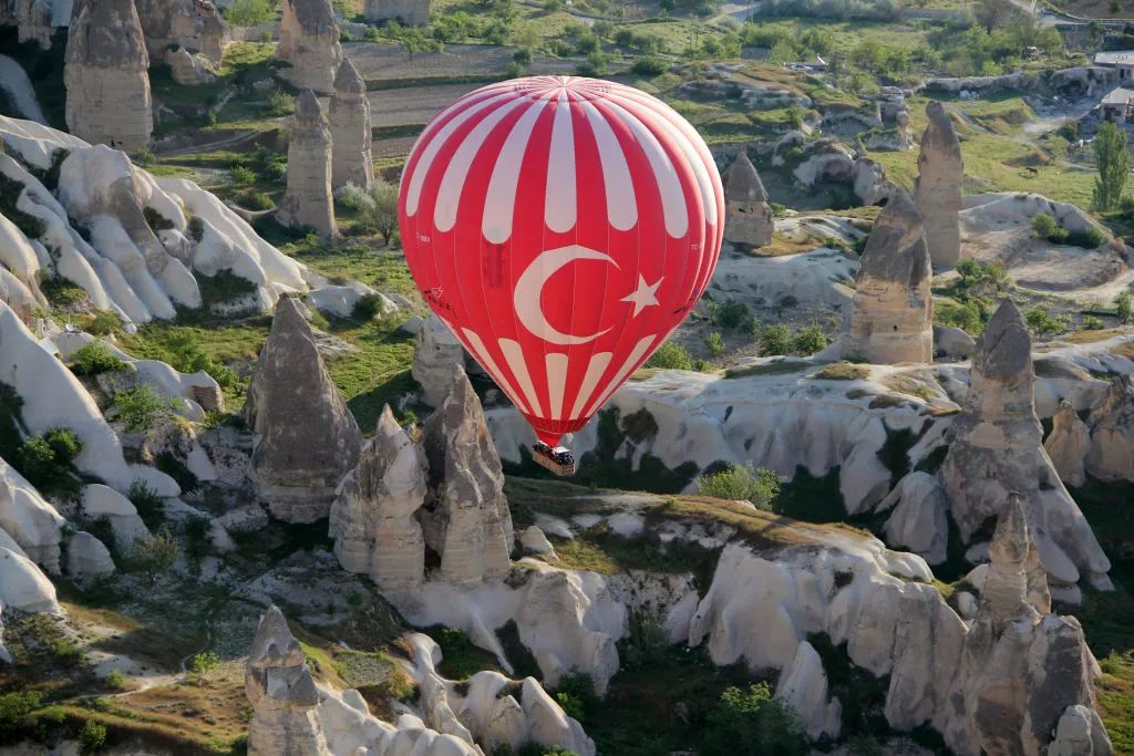 A hot air balloon with the Turkish flag sails past fairy chimneys, a rock formation typical for Cappadocia. 