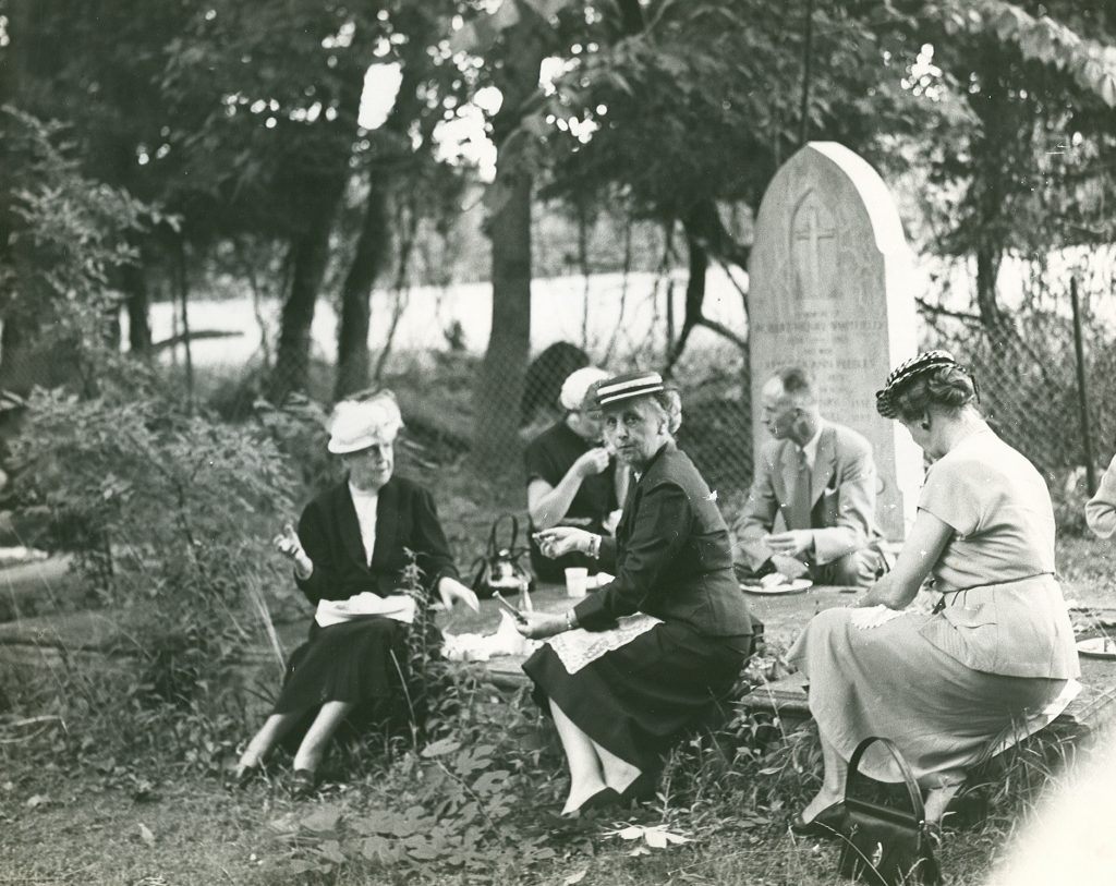 A small group picnics on ledger-style tombstones in Historic St. Luke's Ancient Cemetery. The photo is not dated but is believed to have been taken prior to St. Luke's 1957 Pilgrimage Service.