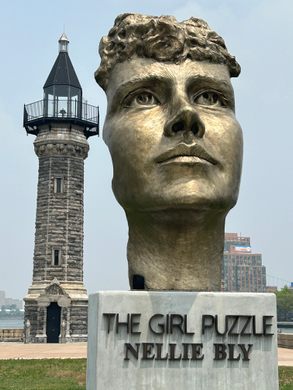 A bronze fast of a female face atop a pedestal with a lighthouse in the background