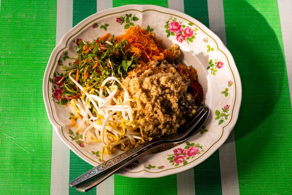 In this salad, rice gets topped with pinches of toasted coconut, dried fish, finely-sliced herbs and vegetables, bean sprouts, a tiny knot of noodles, crushed black pepper and dried chili. 