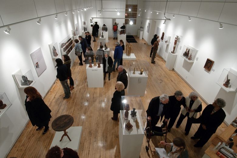 Visitors view sculptures in glass cases and on walls within a brightly lit art gallery with wood floors.