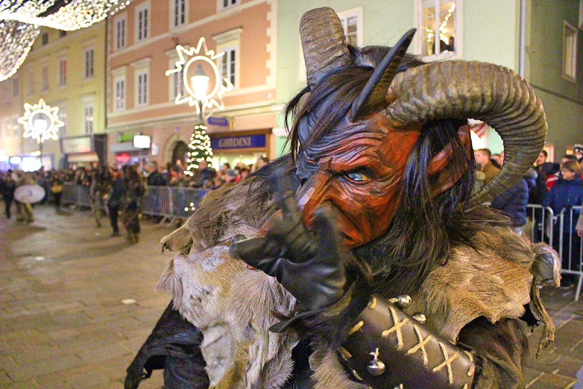 A Krampus waves while wearing a traditional wooden mask and animal pelts around his shoulders.