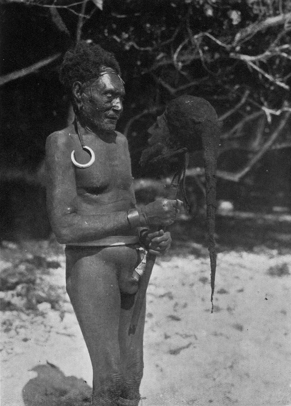 An image from the 1934 book <em>Malekula: A Vanishing People in the New Hebrides</em>, showing a man holding a cured head wearing a spider web cap.