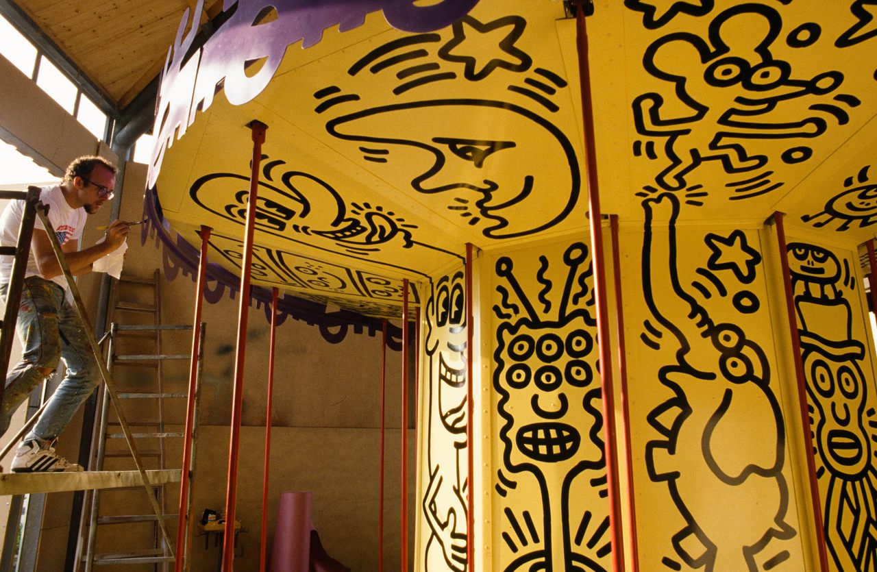 在这张未注明日期的照片中，美国艺术家基思·哈林正在他为1987年的“露娜·露娜”设计的彩色旋转木马上工作。旋转木马的特点是基于他成名的线条画人物的三维人物。
