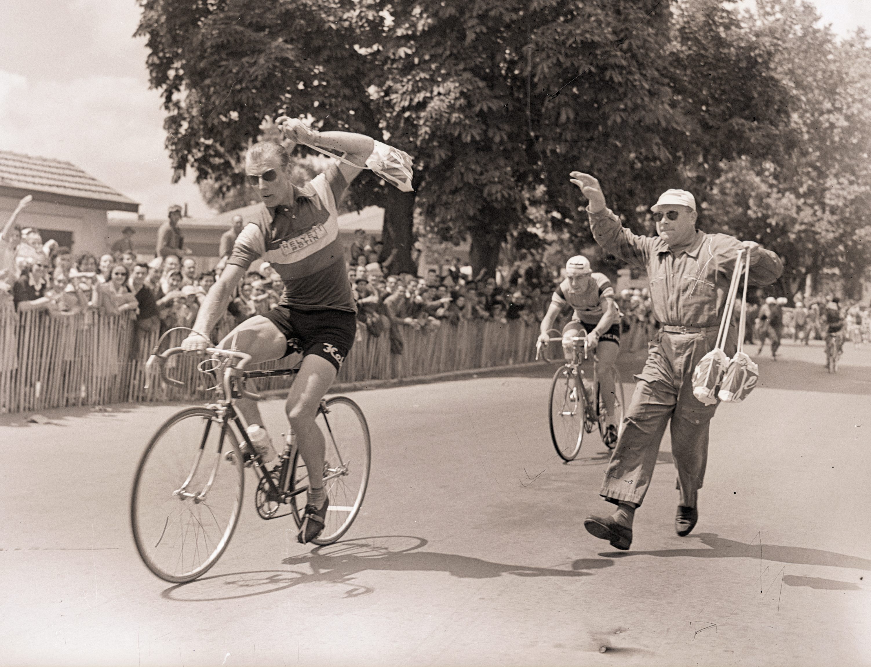 A soigneur performs a successful musette handoff to cyclist Andre Darrigade in 1956. 