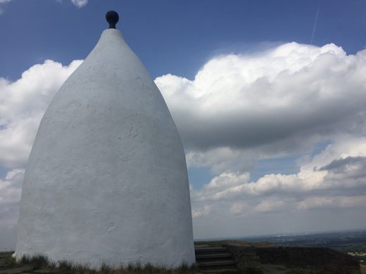 White conical shaped structure with a black bulb-like pinnacle with clouds in the background.