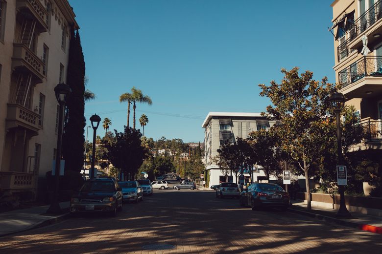 Parked cars line a paved street between apartment buildings with palm trees and the Hollywood sign in the distance.