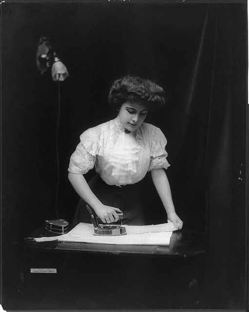 A woman uses an electric iron plugged into light socket, c. 1908.