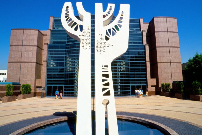 A tall white abstract sculpture stands in a fountain plaza before the glass and stone facade of the Museum of Tolerance.