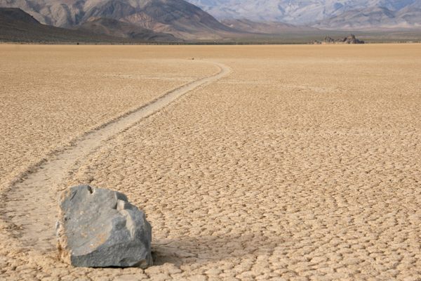 A medium gray rock on the desert floor, with a trail in the dirt behind it showing how the rock has moved.