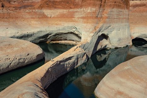 A 171-foot-tall white stain, left by the retreating waters of Lake Powell, looms over a lone human as he strides across a sandstone fin in the heart of Glen Canyon.