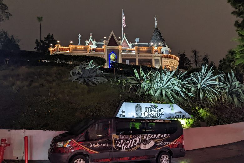 The Magic Castle, a Victorian-style mansion with turrets and lights, sits on a hill above a branded van at night.